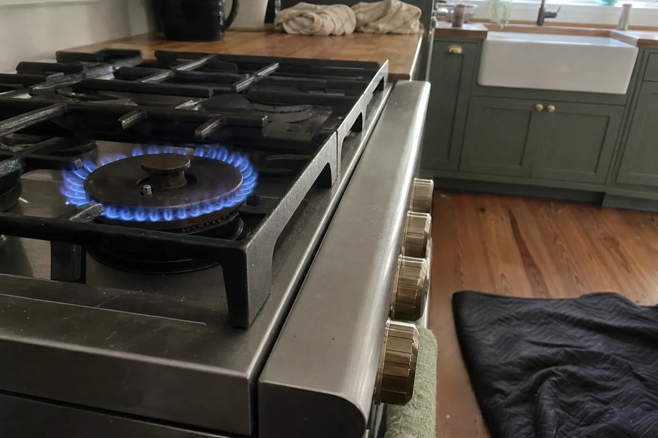 One gas burner burning in a steady ring of blue flame beneath a heavy black cast-iron grate, brass control knobs along the stainless front edge, with sage-green cabinets and a white farmhouse sink behind